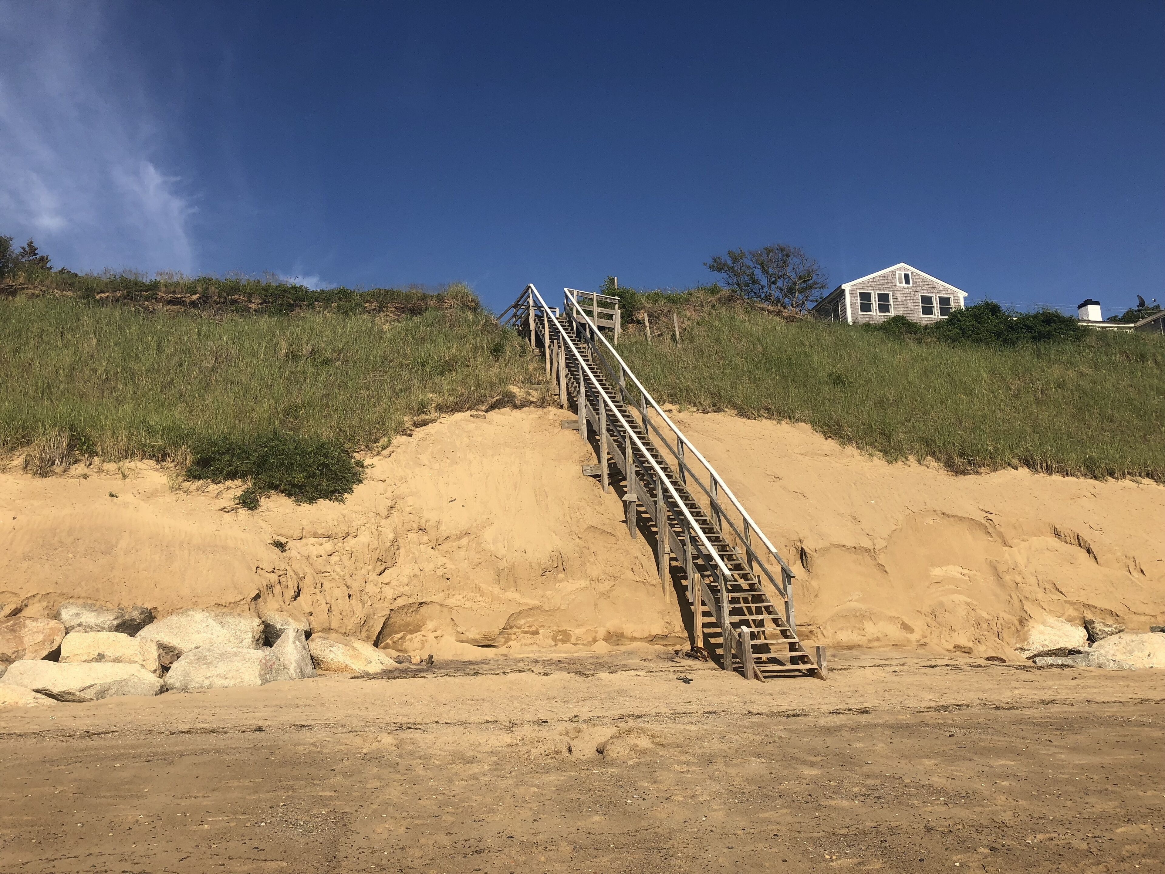 Una playa arenosa con una escalera de madera que sube una duna, una casa blanca al fondo y un cielo azul despejado.