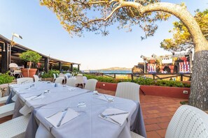Un área de comedor al aire libre con mesas dispuestas para una comida, con vistas a la playa y al mar.