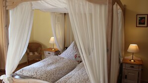 A four-poster canopy bed with a floral bedspread and pillows, a wooden nightstand with a lamp, and a wicker chair in a room with yellow walls. - Apartment at the castle Eltz (Wierschem)