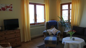 A living room with a television on a wooden dresser, a blue armchair with a white pillow, a round table with a vase of flowers, and a dark-colored sofa. - Apartment at the castle Eltz (Wierschem)