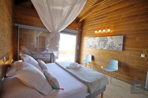 A bedroom with a bed, two chairs, a window with curtains, and a wall-mounted light fixture.