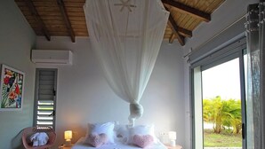 A bedroom with a canopy bed, a pink chair, a bedside table with a lamp, and a view of palm trees outside.