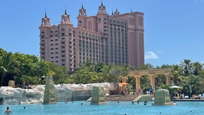 A large swimming pool with people swimming and sunbathing, surrounded by palm trees and a large hotel building in the background. - 3 Bedroom Lockoff  Harborside Atlantis April 19-26, 2025 with 10 Wristbands (Nassau)
