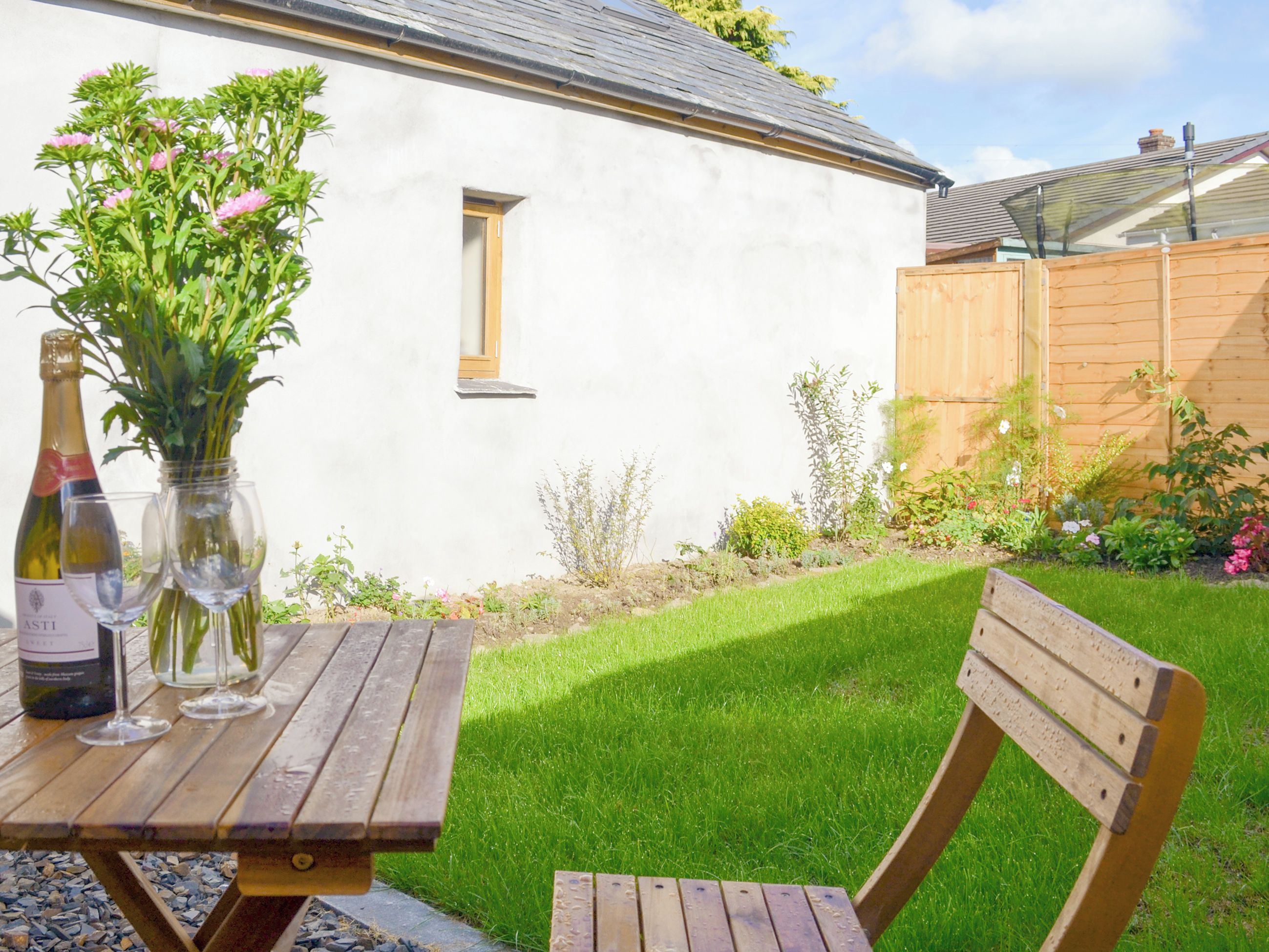 Une table en bois avec une bouteille et des verres, un bâtiment blanc, une clôture en bois et une cour couverte d’herbe.