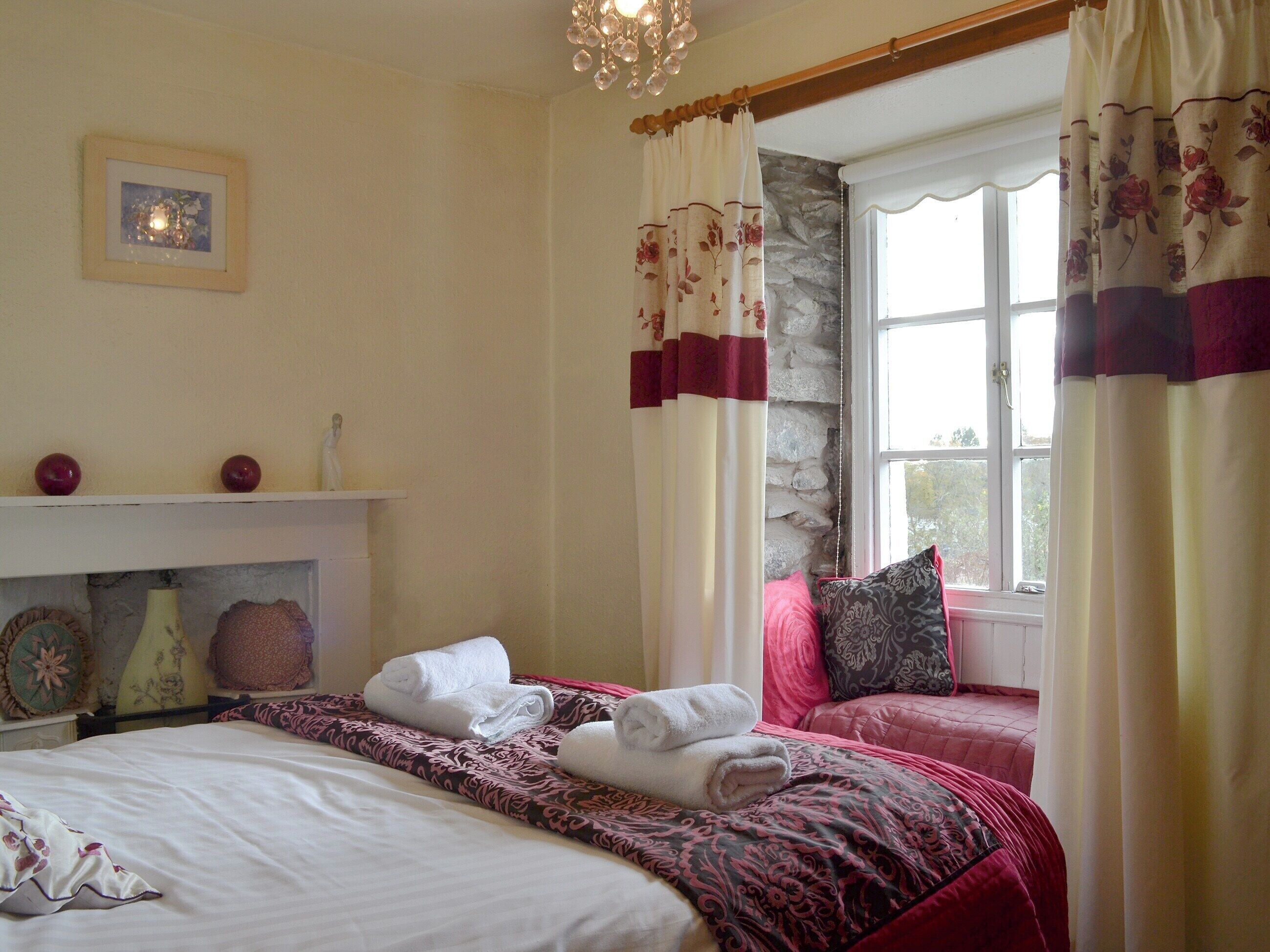 A bedroom with a bed, a window with curtains, a stone wall, and a chandelier.