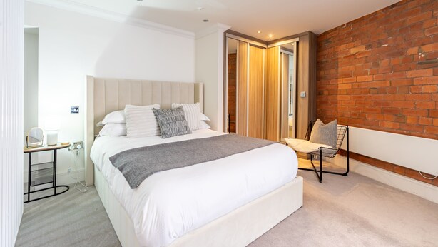 A modern bedroom with a large bed, a bedside table, a chair, and a brick wall. - Cocoa Loft - York city centre studio (with parking) (York)