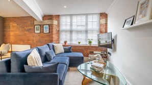 A modern living room with a blue sectional sofa, a glass coffee table, and a brick wall. - Cocoa Loft - York city centre studio (with parking) (York)