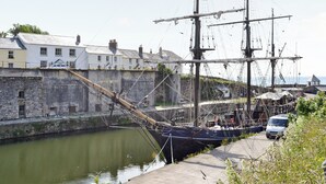 Een historisch schip afgemeerd in een haven, met een stenen muur en gebouwen op de achtergrond.