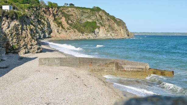 Een rotsachtig kustlandschap met een kiezelstrand, een klif met schaarse begroeiing en de zee.