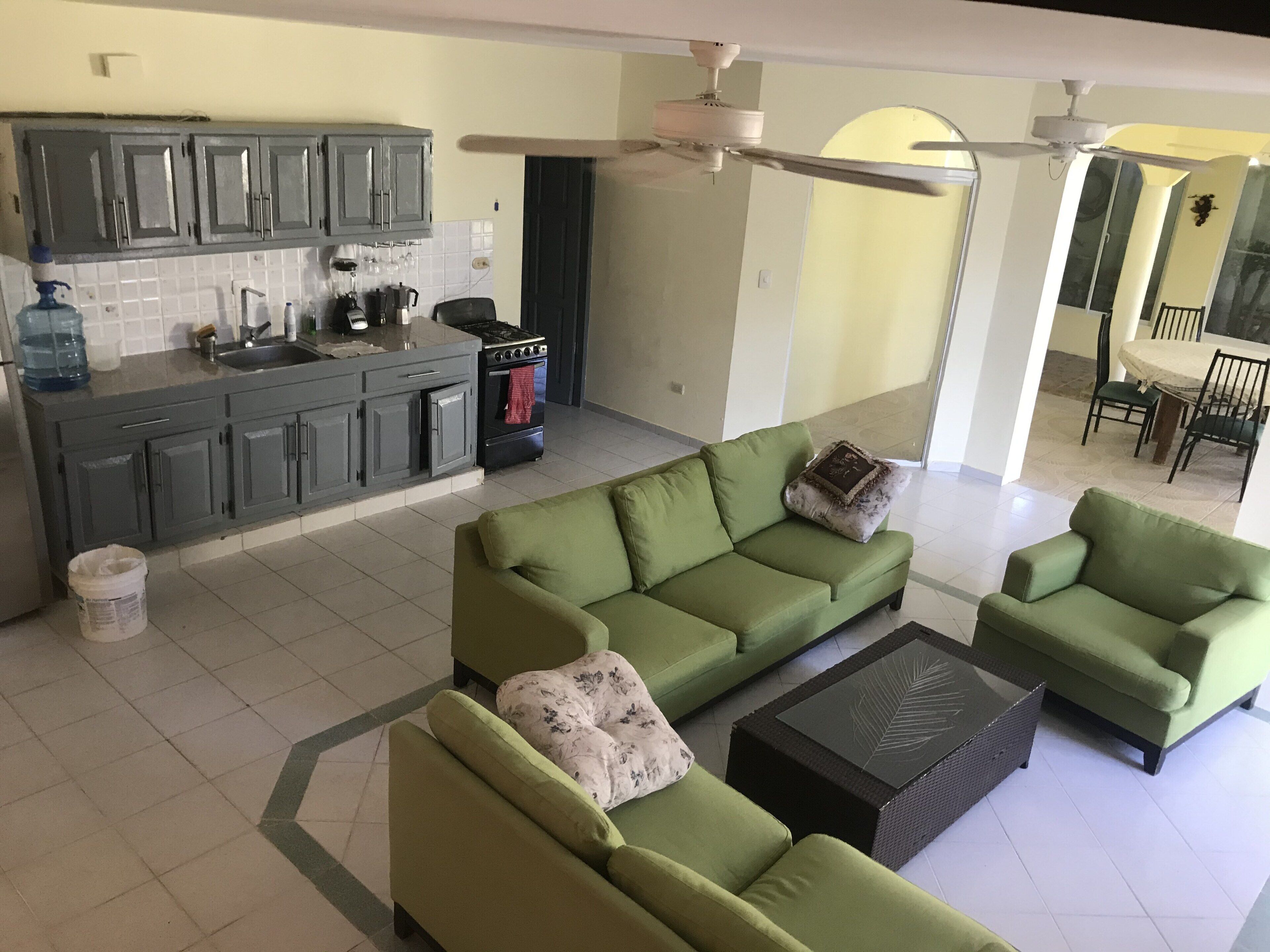 Amplia sala de estar con un juego de sofá verde, una mesa de centro y una cocina con gabinetes grises y una estufa.