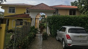 A yellow house with a red-tiled roof, a gated entrance, and a parked silver car. - Three bedroom apartment with private pool (Sosua)
