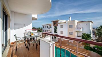 A balcony with a pool, a dining table with chairs, and a view of neighboring buildings.