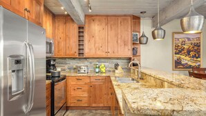 Una cocina con gabinetes de madera, un refrigerador de acero inoxidable, encimeras de granito y una lámpara colgante.