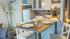 A dining area with a wooden table set for four, white chairs, and a kitchen in the background with white cabinets and a hanging light fixture. - Apartments Harmony 18 (Belgrade)