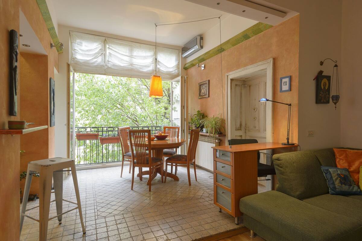 A cozy living room with a green sofa, wooden dining table, and chairs. There's a small stool and a radiator on the floor.