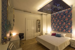 A bedroom with a bed, a desk, a chair, a nightstand, and a wall with a floral pattern.