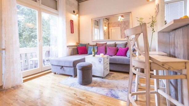A modern living room with a sofa, ottoman, and a wooden chair. There is a large window providing natural light and a view of greenery outside. - Apartments Harmony 18 (Belgrade)