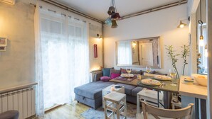 A cozy living room with a grey sofa, a dining table set for two, and a small kitchen area. - Apartments Harmony 18 (Belgrade)