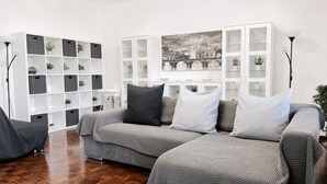 A modern living room with a grey sofa set, a white shelving unit with plants, and a large framed cityscape picture on the wall.