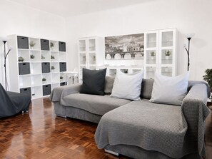 A modern living room with a grey sofa set, a white shelving unit with plants, and a large framed cityscape picture on the wall.