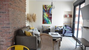 Una sala de estar moderna con una pared de ladrillo, un cuadro abstracto y colorido, un sofá con cojines estampados, un sillón de mimbre, una mesa de centro y una estantería con libros y adornos.