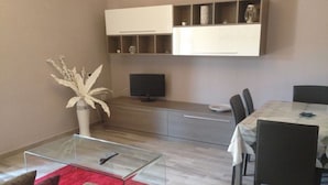 A modern living room with a glass coffee table, a red rug, a sofa, a dining table with chairs, a flat-screen TV, and built-in shelving above the TV. - Central Apartments Ortensia (Caulonia Marina)