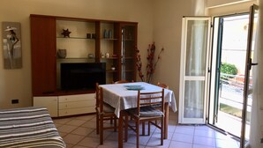 A compact living space with a dining table, chairs, a TV, and a sliding glass door leading to a balcony. - Central Apartments Ortensia (Caulonia Marina)