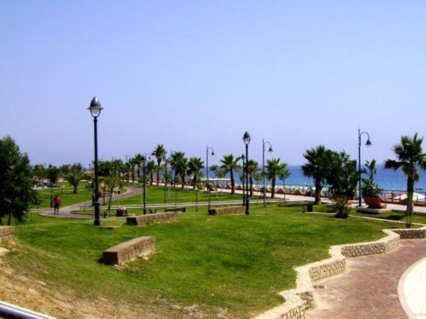 A coastal promenade with a paved walkway, palm trees, and street lamps.