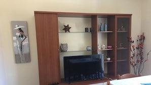 A living room with a TV on a wooden cabinet, a glass shelf with decorative items, and a painting on the wall. - Central Apartments Ortensia (Caulonia Marina)