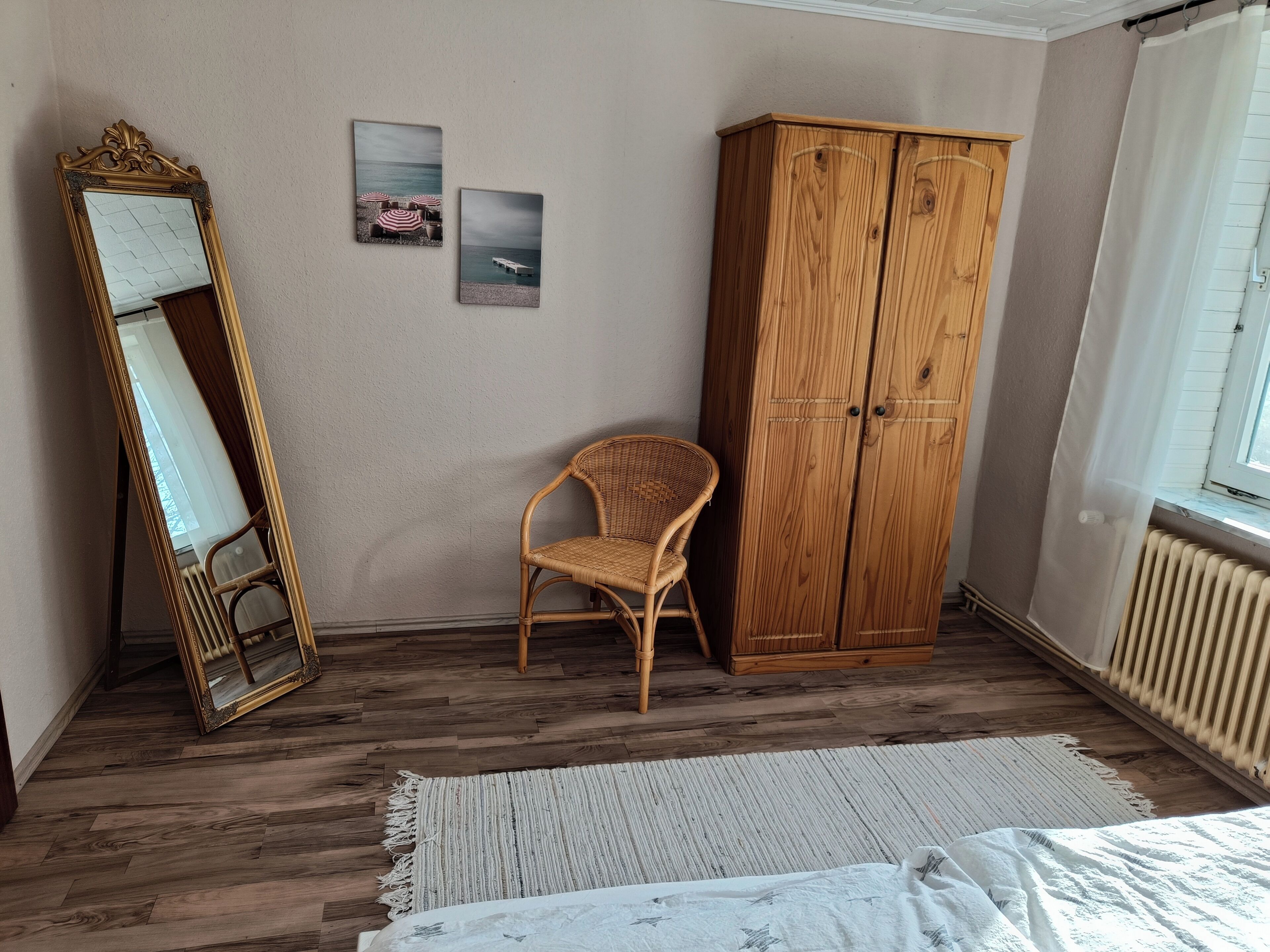 A room with a wooden floor, a wicker chair, a wooden wardrobe, a mirror, and two framed pictures on the wall.