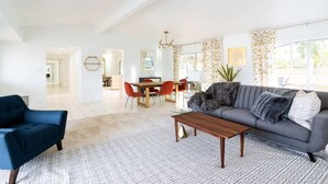 Uma sala de estar ampla, com um sofá cinzento, uma poltrona azul e uma mesa de centro de madeira. Ao fundo, há uma área de refeições com uma mesa e cadeiras.