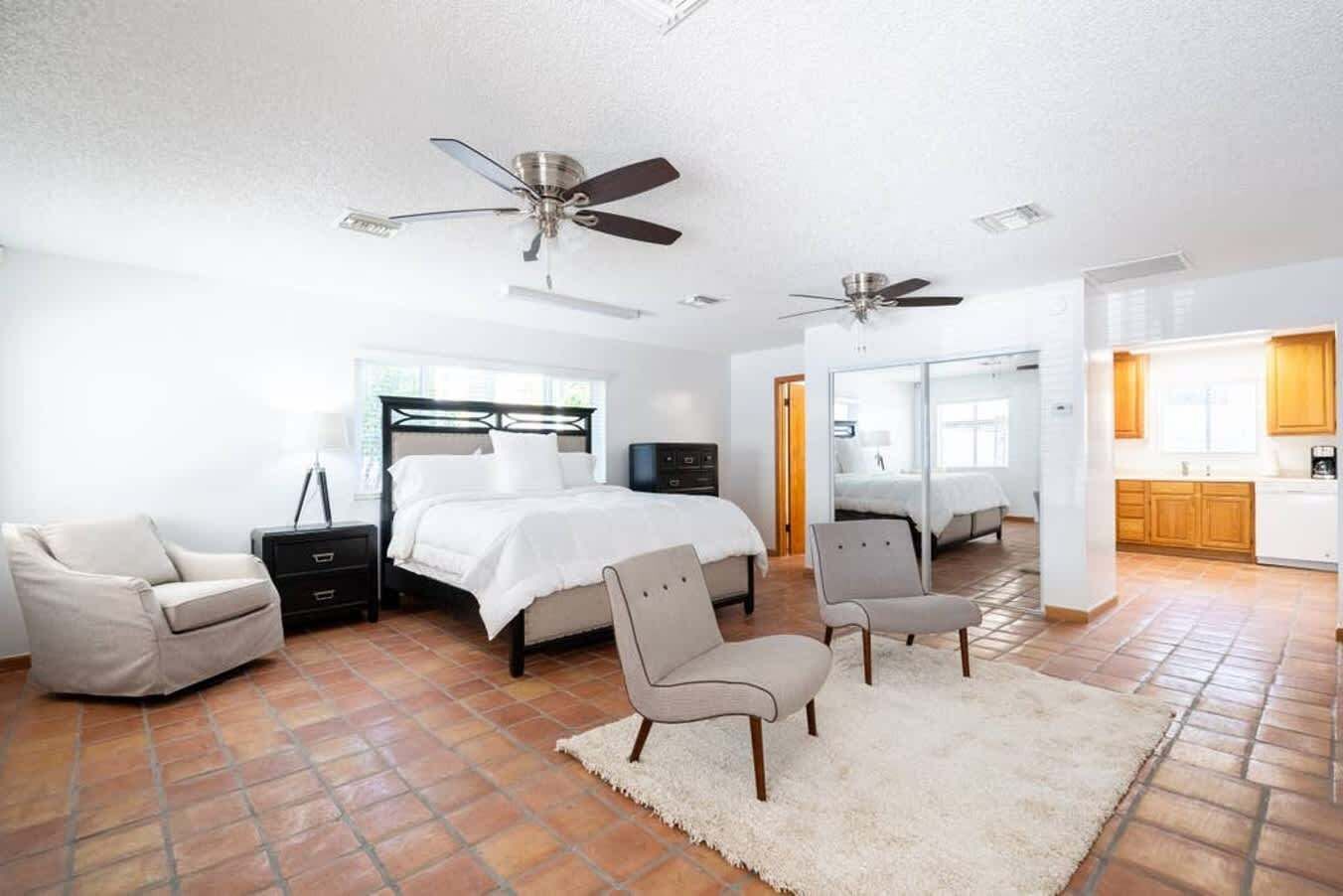 Une chambre spacieuse avec un grand lit, deux fauteuils, une table de chevet, un bureau, un ventilateur de plafond et une vue sur la cuisine adjacente.