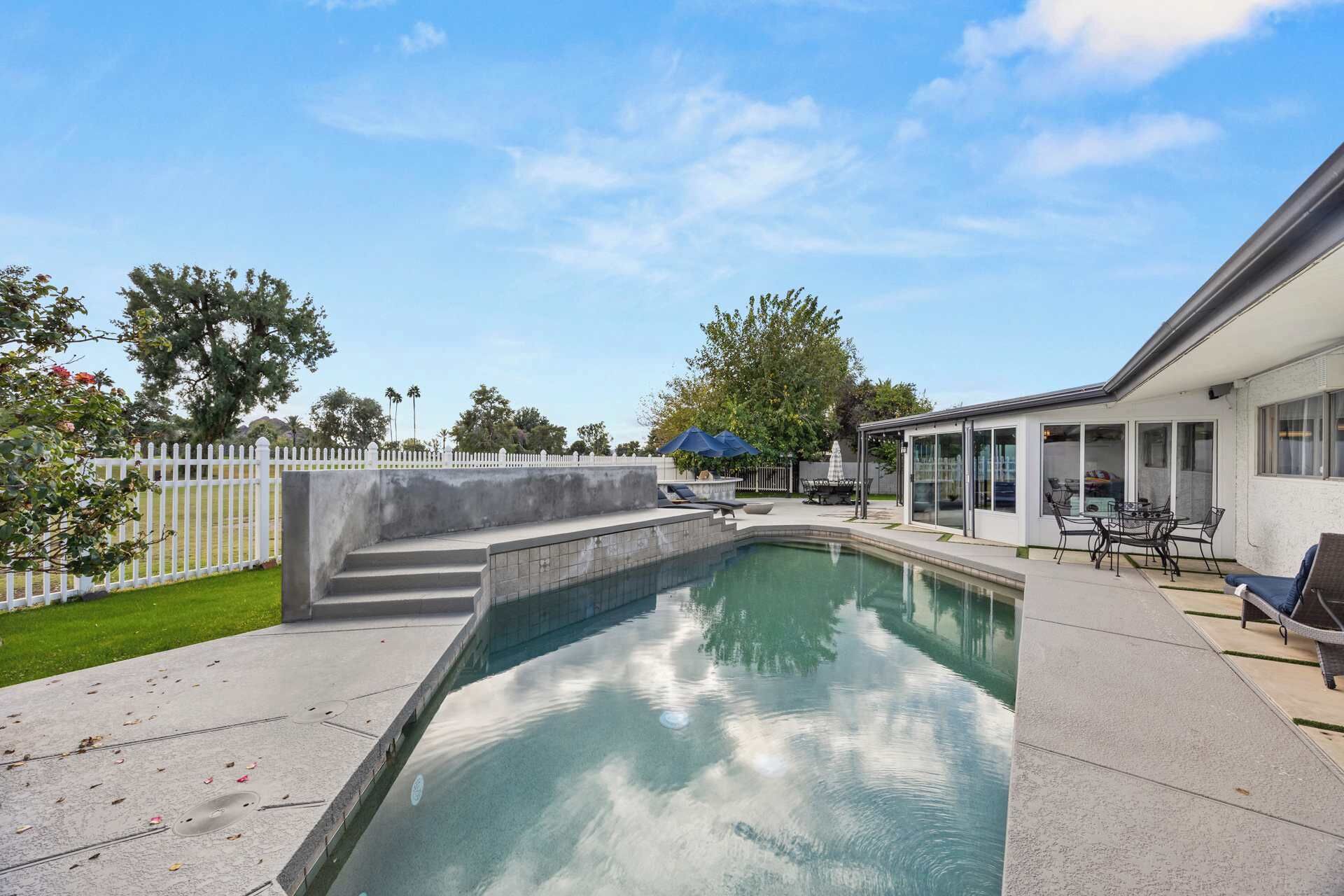 A swimming pool with a concrete deck, a covered patio area with outdoor furniture, and a white fence.