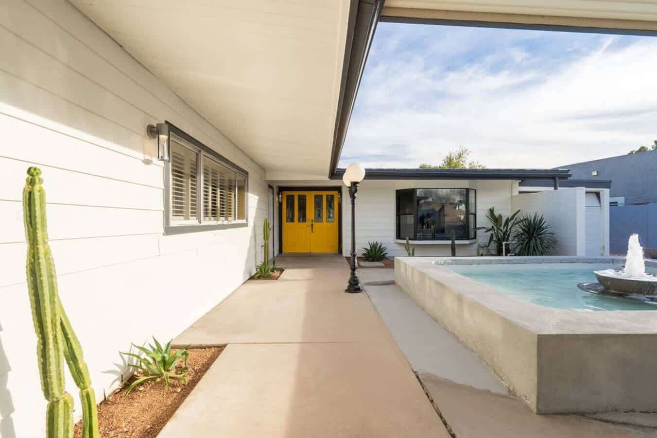 Une maison moderne avec une porte jaune, une petite piscine avec une fontaine et un cactus au premier plan.