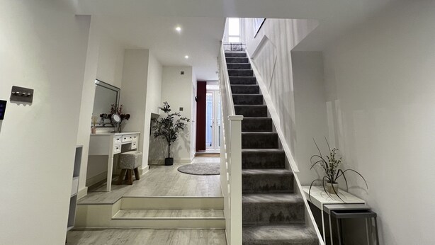 Un intĂ©rieur moderne avec un escalier menant Ă un deuxiĂšme Ă©tage, un couloir dotĂ© dâun plancher de couleur claire et un petit coin salon avec un tabouret et une plante.