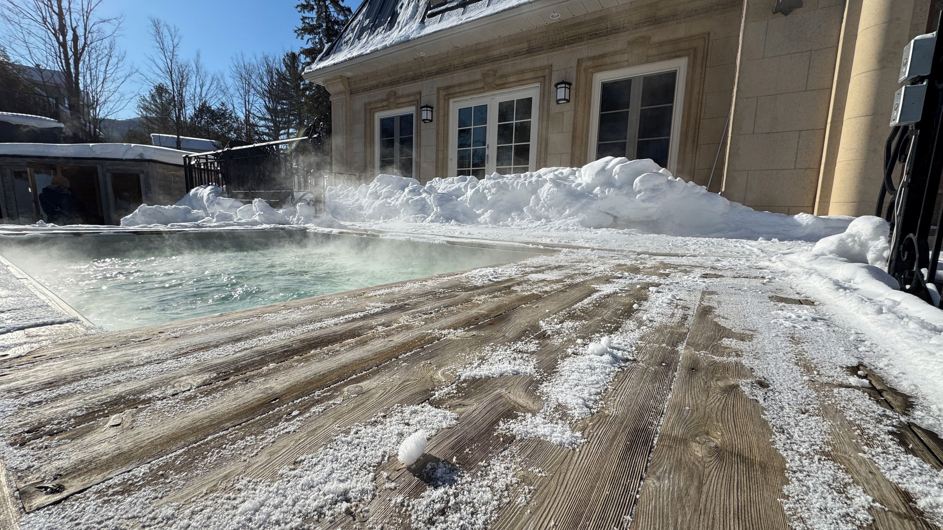Una terraza de madera con nieve, una piscina y un edificio con ventanas y puertas.