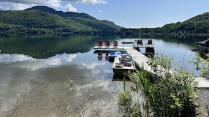 Een vredig steiger aan het meer met een rij Adirondack-stoelen, een klein bootje en een houten pier die zich uitstrekt over kalm water, omringd door weelderige groene heuvels.