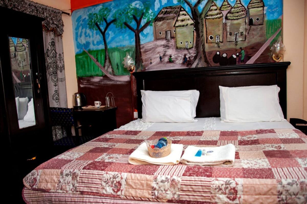 A neatly made bed with a quilt, a small table with a bottle and a cup, and a painting of houses on the wall.