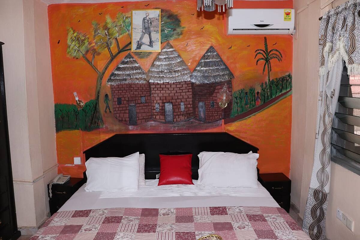A bedroom with a bed, a red pillow, a nightstand, and a wall mural depicting a building and trees.