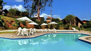 A swimming pool with white lounge chairs and umbrellas, surrounded by greenery and buildings. - House in the Mountains - Exclusive view! (Gravatá)