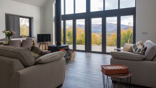 A spacious living room with a large window offering a mountain view, a fireplace, and a cozy seating area. - Relax and Refuel to the Rhythm of Nature at Fia Lodge. (Glencar)
