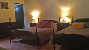 Una habitación con dos camas de madera, cada una con una mesita de noche y una lámpara. Se ve una puerta que da a otra habitación y una pequeña mesa con una lámpara.