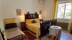 Un dormitorio con una cama, dos mesitas de noche, una silla, una mesa redonda y un ventanal con cortinas.