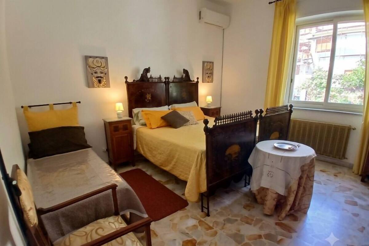Un dormitorio con una cama, dos mesitas de noche, una silla, una mesa redonda y un ventanal con cortinas.