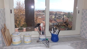 Una cocina con un lavamanos, una ventana que ofrece vistas a un paisaje urbano, y diferentes utensilios y recipientes sobre la encimera.