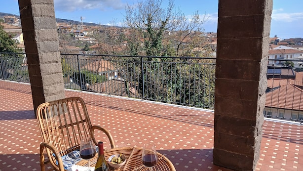 Un balcón con mobiliario de mimbre, una botella de vino y copas, con vista a un barrio residencial y cielo despejado.