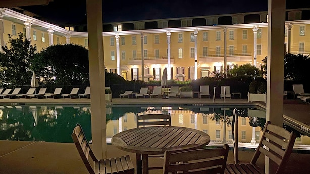 Un espace aménagé au bord de la piscine, avec des chaises longues et une table, dans lequel se reflète le bâtiment situé à l’arrière-plan.