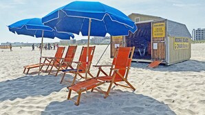 Des chaises de plage et des parasols se trouvent à proximité d’une petite structure en bois portant l’inscription « LOCATION DE PLANCHES DE SURF ET DE CHAISES ».