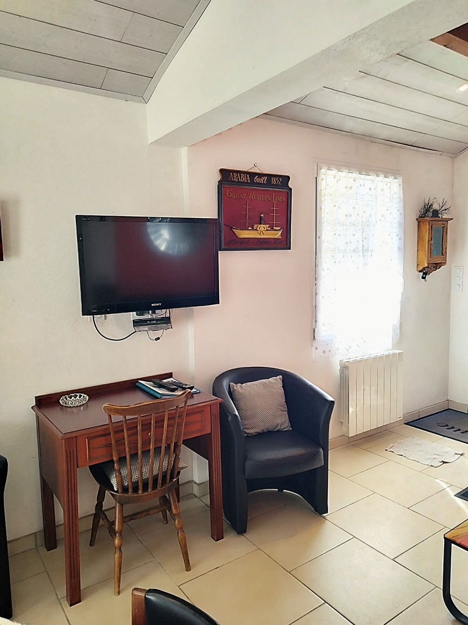 A room with a flat-screen TV mounted on the wall, a wooden desk with a chair, a black armchair, and a window with a decorative curtain.
