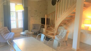 A living room with a white sofa, a round coffee table, and a TV on a wooden stand. There is a staircase with a wooden railing leading to an upper level. - LA CARRETERIE - "Le Philosophe" (cottage 4 pers. 70m2) (Pineuilh)